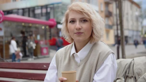Молода блондинка бізнес-леді в бежевому жилеті і біла блузка сидить на лавці в центрі міста і посміхається з кавою в руці — стокове відео