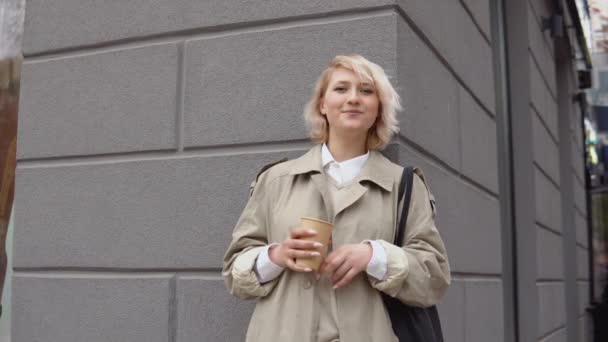 Молода блондинка бізнес-леді в бежевому траншеї пальто і біла блузка з чорною сумкою і витягнути каву в її руках стоїть, спираючись на стіну будівлі і дивиться на камеру — стокове відео