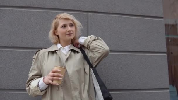 Joven mujer de negocios rubia en una gabardina beige y blusa blanca con una bolsa negra y café para llevar en sus manos se para cerca de la pared gris del edificio y mira a la cámara — Vídeos de Stock