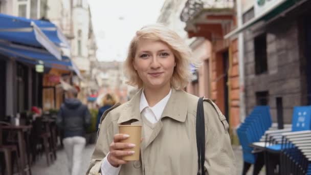 Jonge blonde zakenvrouw in een beige trench jas en witte blouse met een zwarte tas en afhaalkoffie in haar handen staat op een gezellige straat met restaurants en kijkt naar de camera — Stockvideo