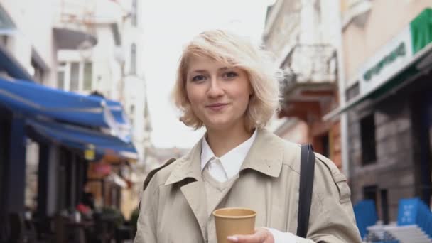 Νεαρή επιχειρηματίας με μπεζ καμπαρντίνα και λευκή μπλούζα με ένα φλιτζάνι καφέ και ένα κινητό τηλέφωνο στα χέρια της κοιτάζει την κάμερα ενώ στέκεται στο δρόμο. Πορτρέτο μιας χαμογελαστής νεαρής γυναίκας — Αρχείο Βίντεο