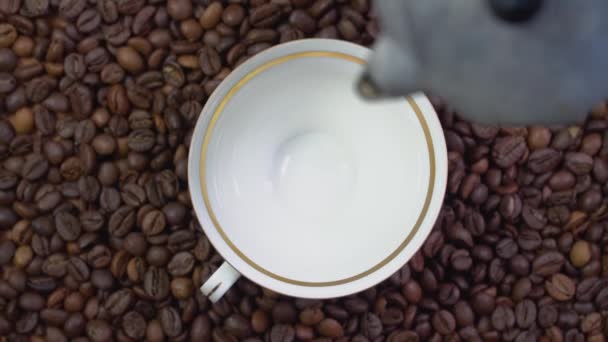 En una taza de porcelana con dorado vierta el café de una cafetera. Taza con café sobre un fondo de granos de café. Vista superior — Vídeo de stock