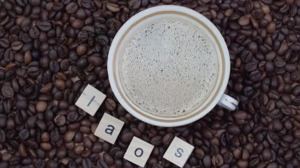 Κάτοψη ενός φλιτζανιού καφέ σε φόντο κόκκων καφέ με την επιγραφή Λάος. Χώρα παραγωγής καφέ — Αρχείο Βίντεο