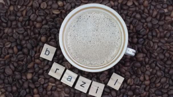 Вид сверху на чашку кофе на фоне кофейных зерен с надписью Бразилия. Страна производства кофе — стоковое видео