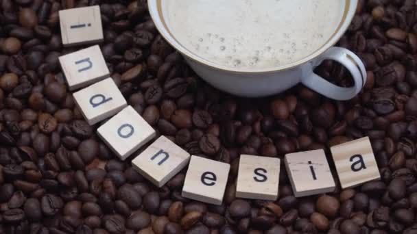 Πορσελάνη και επίχρυσο φλιτζάνι καφέ σε φόντο κόκκων καφέ με την επιγραφή Ινδονησία. Χώρα παραγωγής καφέ — Αρχείο Βίντεο