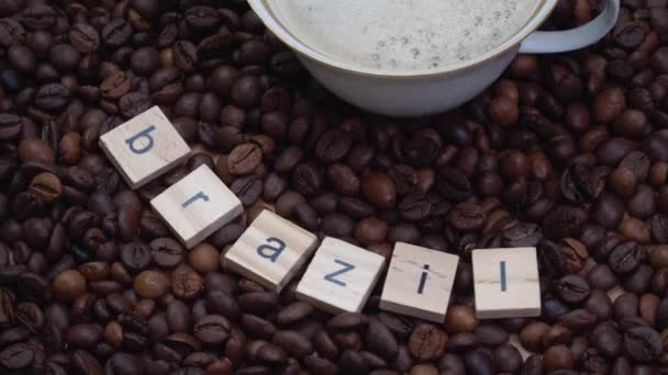 Кофейные зерна с надписью Бразилия и фарфоровая чашка с кофе на них. Производство, экспорт и импорт кофе — стоковое видео
