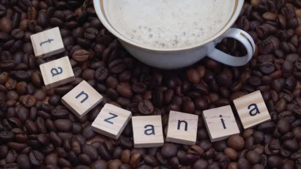 Κόκκοι καφέ με την επιγραφή Τανζανία και ένα φλιτζάνι πορσελάνη με καφέ πάνω τους. Χώρα παραγωγής καφέ — Αρχείο Βίντεο