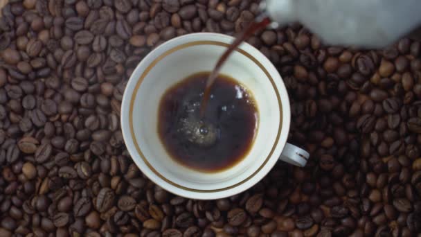 En una taza de porcelana con dorado vierta el café de una cafetera. Taza con café sobre un fondo de granos de café. Vista superior — Vídeos de Stock