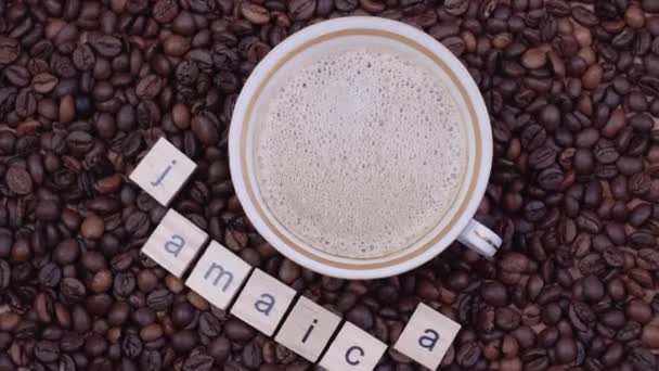 Cup of coffee on a background of coffee beans with the word Jamaica outlined. Production, export and import of coffee. Top view — Stock Video