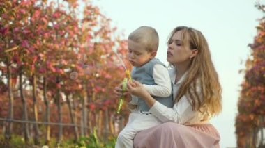 Oğluyla annesinin eğlencesine. Oğlu bahçede pembe çiçeklerle sabun köpükleri yapıyor. Ebeveynler ve çocuklar arasındaki nazik ilişki