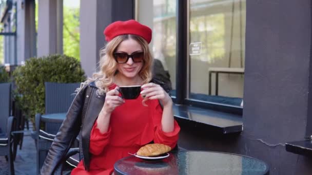 Stilvoller Kaffee. Die Blondine trinkt Kaffee mit einem Croissant. Moderne schwarze Terrasse des Restaurants. Junge Frau mit Sonnenbrille und Baskenmütze — Stockvideo