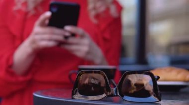 Kruvasanla kahve molası. Restoran terasında cep telefonuyla çalışan modern şehirli genç bir kadın. Kesintisiz internet erişimi. Güneş gözlüklerinin görüntüsünü kapat