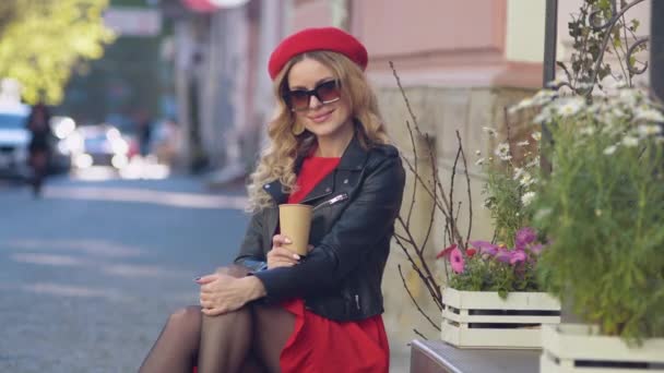 Молодая красивая женщина в красном берете и платье пьет кофе и смотрит в камеру. Фотосессия на улице — стоковое видео