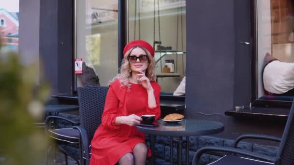 Estética matutina lenta.Jovem mulher atraente romântico sorrindo olhando para a câmera. Mulher de vestido vermelho e boina sentada com uma xícara de café em uma mesa na rua — Vídeo de Stock