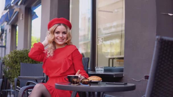 Jovem mulher atraente romântico sorrindo olhando para a câmera. Mulher de vestido vermelho e boina sentada com uma xícara de café em uma mesa na rua — Vídeo de Stock