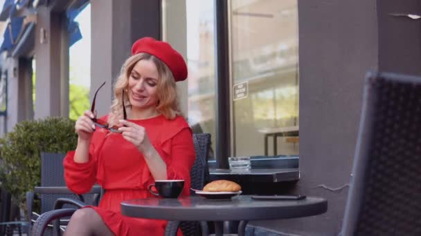 Mladá žena se usmívá a nosí stylové sluneční brýle. Žena v červených šatech a baretu sedící s šálkem kávy a croissantem na stole na ulici — Stock video