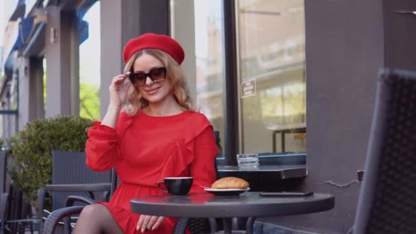 Молодая женщина улыбается и настраивает стильные солнечные очки. Женщина в красном платье и берете сидит с чашкой кофе и круассаном на столе на улице — стоковое видео
