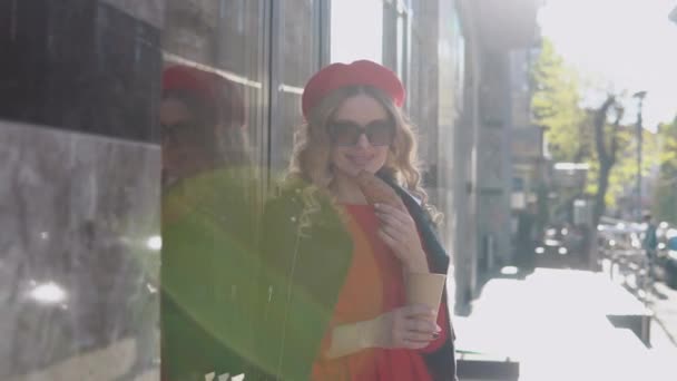 Loira jovem elegante em um vestido vermelho e trança preta fica em uma rua acolhedora ao sol da manhã. Café da manhã com croissant — Vídeo de Stock