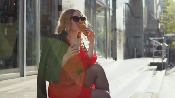 Elegante belleza bebe café y come un croissant sentado en las escaleras a la entrada de un edificio moderno. Estilo de vida empresarial, gestión del tiempo, recreación de una mujer moderna — Vídeos de Stock