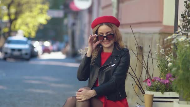 Молодая красивая женщина в красном берете и платье надевает очки и смотрит в камеру. Фотосессия на улице — стоковое видео