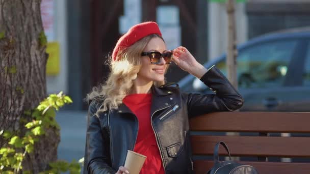 Joven mujer hermosa en una boina roja y vestido, gafas negras y una trenza sentada en un banco del parque. Sesión de fotos en la calle — Vídeo de stock