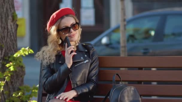 Mladá krásná žena v červeném baretu a šatech, černé brýle a cop sedí na lavičce v parku. Buněčná komunikace jako životní styl. Přestávka na oběd uprostřed pracovního dne — Stock video