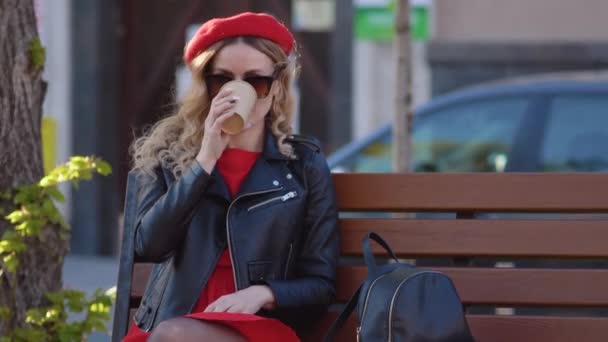 Молодая красивая женщина в красном берет и платье, черные очки и коса сидит на скамейке в парке с чашкой кофе. Перерыв на обед в середине рабочего дня — стоковое видео