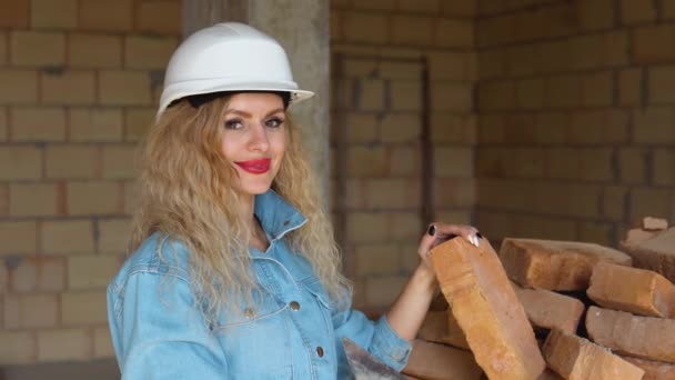 Hermosa mujer con maquillaje y manicura sosteniendo un ladrillo en el sitio de construcción. Mujer con casco y vaqueros de trabajo — Vídeos de Stock