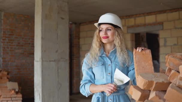 Красивая женщина каменщик с макияжем и маникюром держа кирпич и лопатку на строительной площадке. Женщина-мастер в шлеме и джинсовой рабочей форме — стоковое видео