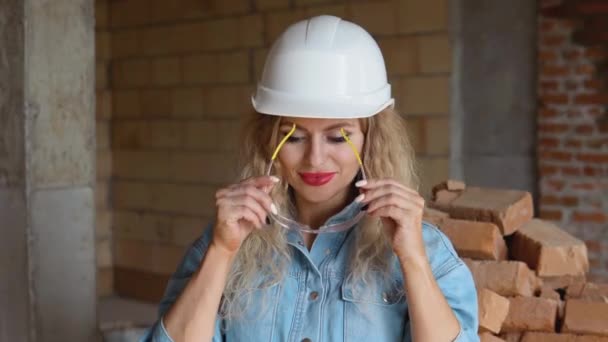 Mujer joven en un casco de trabajo y ropa de mezclilla lleva gafas en el sitio de construcción. Trabajadora en la industria de la construcción — Vídeos de Stock