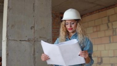 Mimari inşaat planı. Beyaz iş kaskı ve kot pantolon ve gözlüklü genç bir kadın şantiyede duruyor ve bir inşaat planı tutuyor..