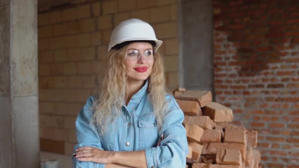 Een jonge vrouw met een werkhelm en denim kleding en bril staat op de bouwplaats. Controle van de veiligheid op de bouwplaats — Stockvideo