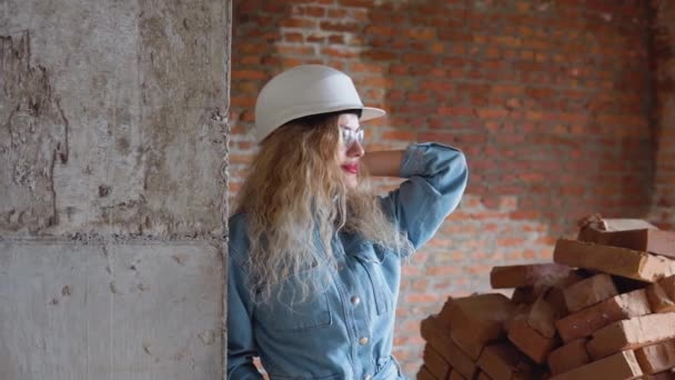 Una mujer joven en un casco de trabajo y ropa de mezclilla y gafas de pie en el sitio de construcción. Estética de las profesiones en activo — Vídeos de Stock