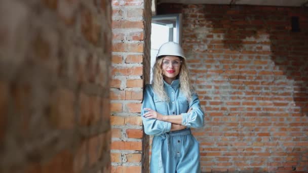 Le processus de construction d'une maison de rêve. Une jeune femme portant un casque de travail et des vêtements et lunettes en denim se tient sur le chantier de construction. — Video