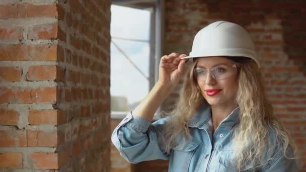 Молодая женщина в белом рабочем шлеме и джинсовой одежде и очках стоит на стройке. Строитель, каменщик, архитектор — стоковое видео