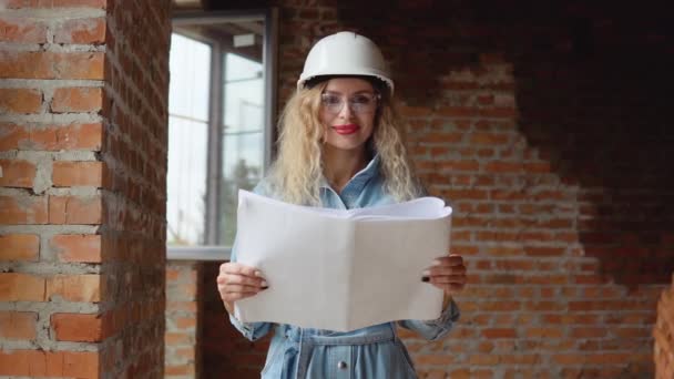 Молодая женщина в белом рабочем шлеме и джинсовой одежде и очках стоит на стройке и держит план строительства. Строитель, каменщик, архитектор — стоковое видео