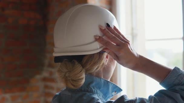 Une femme architecte se tient avec son dos à la caméra et regarde par la fenêtre sur le chantier de construction. Femme constructeur ajuste casque de protection sur sa tête — Video