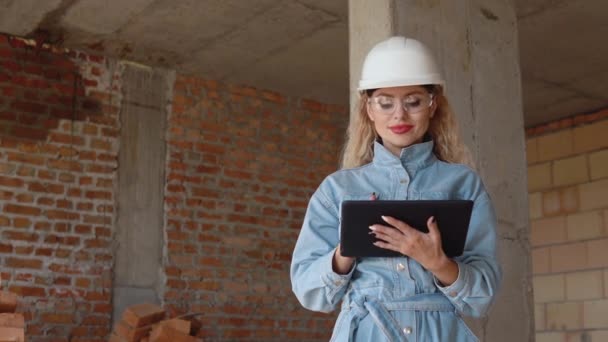 En kvinnlig arkitekt eller murare står i ett nybyggt hus med obehandlade väggar och arbetar på en surfplatta. Modern teknik inom de äldsta yrkena — Stockvideo