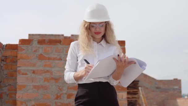 Женщина-архитектор в деловой одежде стоит на улице на строительной площадке. Архитектор делает отметки в архитектурном плане — стоковое видео