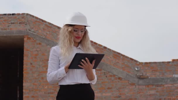 İş kıyafetleri içindeki kadın mimar yeni inşa edilmiş duvarları olmayan bir evde duruyor ve tablet üzerinde çalışıyor. En eski mesleklerdeki modern teknolojiler — Stok video