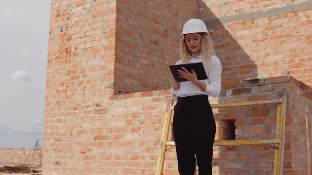 Női építész üzleti öltözékben és egy fehér sisak áll egy újonnan épült ház kezeletlen falak és működik egy tabletta. Modern technológiák a legrégebbi szakmákban — Stock videók