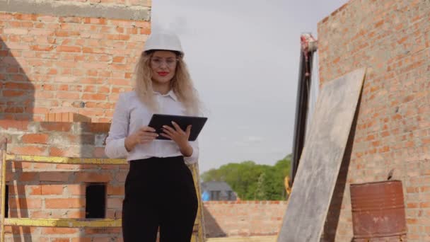 Női építész üzleti öltözékben áll egy újonnan épült ház kezeletlen falak és működik egy tabletta. Modern technológiák a legrégebbi szakmákban — Stock videók