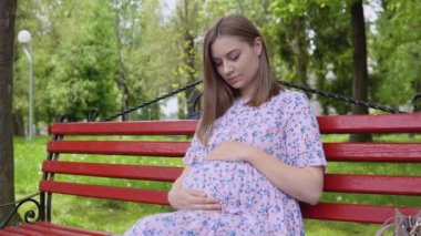 Hamile bir kadın yazın sokakta oturuyor ve ellerini karnının üstüne koyuyor. Hamile bir kadın parkta oturuyor.