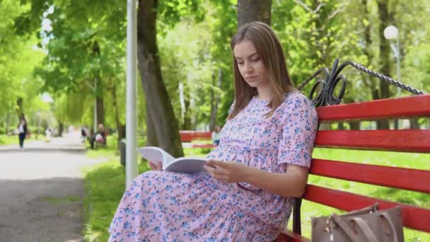 Mujer embarazada en un vestido de verano con un estampado floral pasea por el parque. Una mujer embarazada en un banco del parque lee un libro para las madres embarazadas sobre la lactancia materna. Cuidado del bebé y parto — Vídeos de Stock