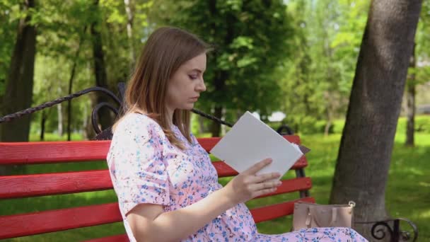 公園を歩く花のプリントの夏のドレスの妊婦。公園のベンチで妊婦が母乳育児についての本を読んでいます。赤ちゃんの世話と出産 — ストック動画