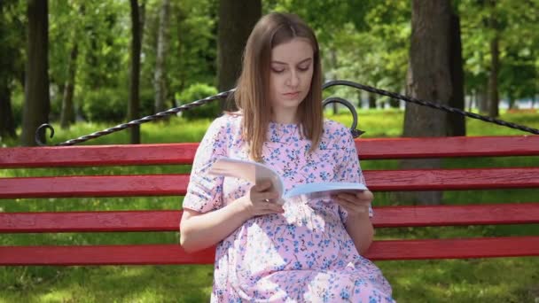 Terhes nő nyári ruhában, virágmintás ruhában sétál a parkban. Egy terhes nő egy padon olvas egy könyvet a várandós anyáknak a szoptatásról. Gyermekgondozás és szülés — Stock videók