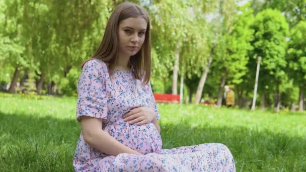 Üzerinde çiçek desenli bir elbiseyle parkta yürüyen hamile bir kadın. Hamile bir kadın parkta çimlerin üzerinde oturmuş karnına sarılıyor. Çocuk doğmadan önce boş zaman. — Stok video