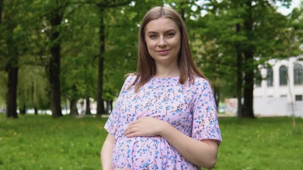 Μια έγκυος γυναίκα με καλοκαιρινό φόρεμα με λουλουδάτο σχέδιο περπατάει στο πάρκο και αναπνέει καθαρό αέρα. Μια έγκυος γυναίκα στέκεται στο δρόμο το καλοκαίρι και κρατά τα χέρια της στην κοιλιά της — Αρχείο Βίντεο