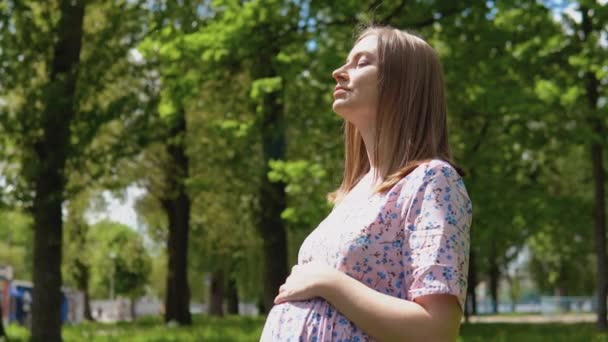 Egy terhes nő nyári ruhában, virágmintás mintával sétál a parkban, és friss levegőt szív. Egy terhes nő áll az utcán nyáron, és tartja a kezét a hasán.. — Stock videók