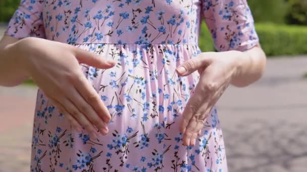 Uma mulher grávida em um vestido de verão com uma estampa floral fica no parque e dobra as mãos em sua barriga com um sinal de coração. Gravidez no verão — Vídeo de Stock